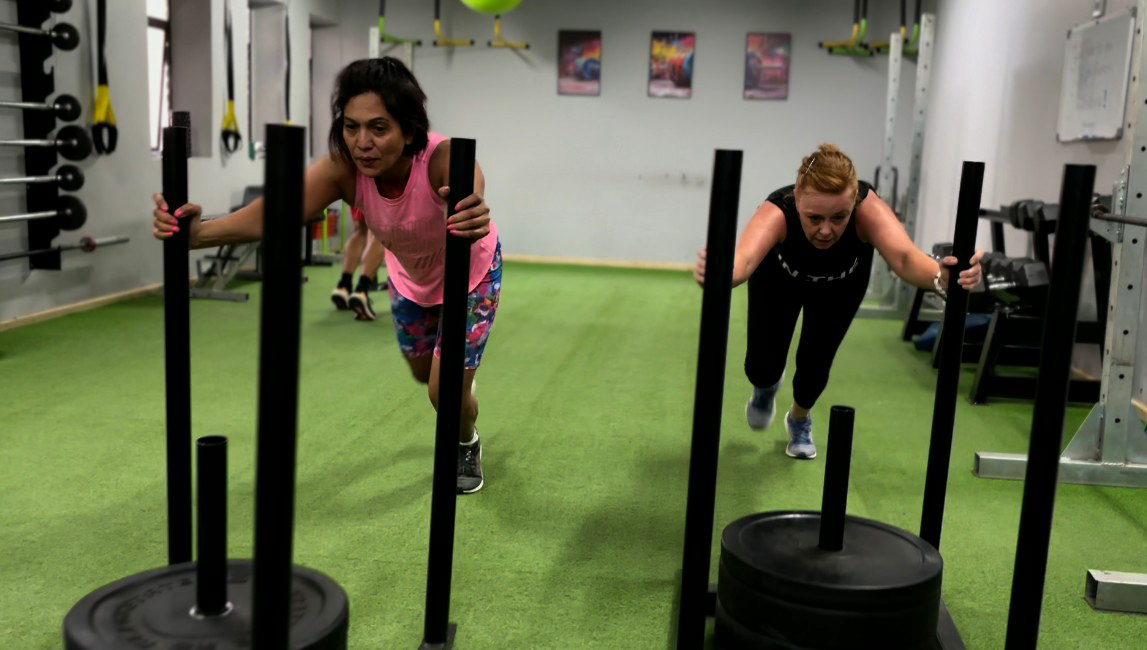 Functional Training: Powering Through the Sled Push at FitRanX Two women pushing weighted sleds across a green turf floor in a FitRanX gym during a functional fitness workout.