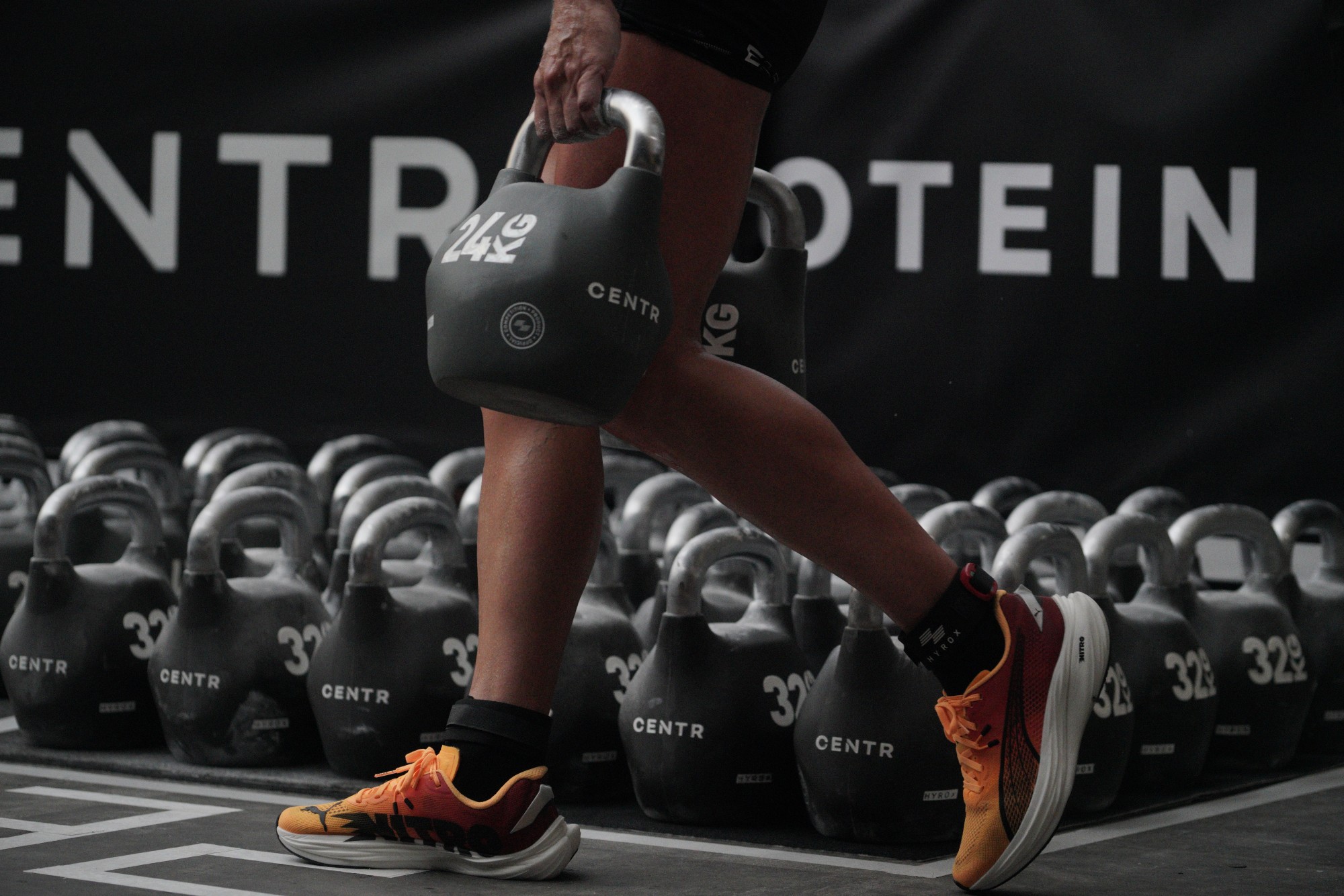 Kettlebell Carry Strength: Core & Grip Training for HYROX Close-up of a person's legs walking while holding a heavy 24kg kettlebell for a Farmer's Carry. A large quantity of other kettlebells is visible in the background, representing the strength and conditioning focus of FitRanX HYROX Training Club in Rivonia.