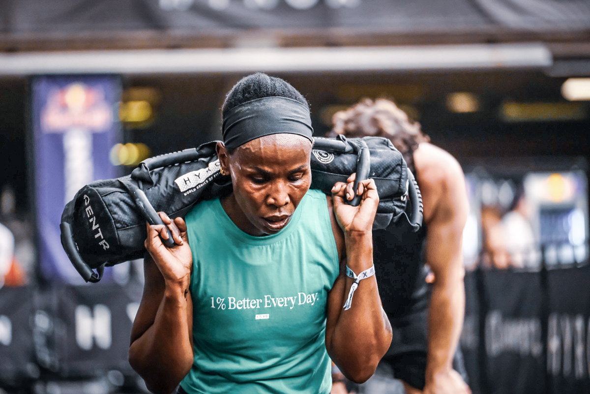 Sandbag Lunges: Building Lower Body Strength for HYROX A determined female athlete performing Sandbag Lunges with a heavy black HYROX sandbag resting on her shoulders. She wears a teal shirt with the text "1% Better Every Day," symbolizing the focus on functional fitness and performance at FitRanX Training Club in Rivonia