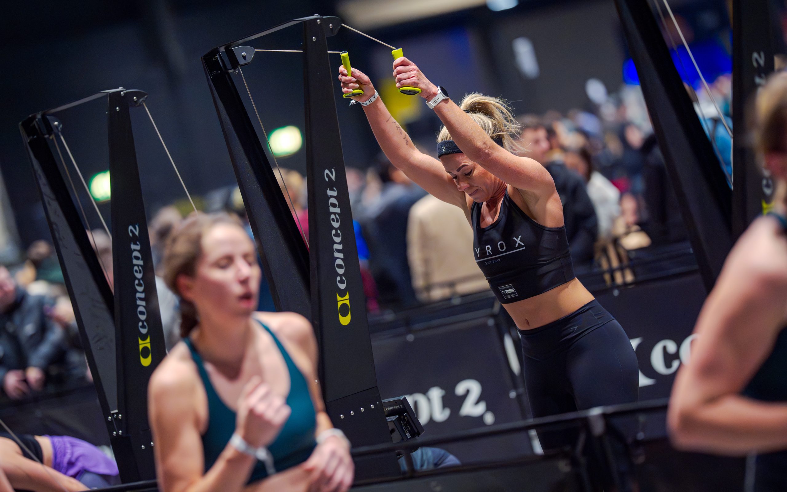 HYROX Training: Female Athlete on the SkiErg at a Fitness Race A female HYROX athlete intensely working the SkiErg station during a race or training. She is wearing a black HYROX crop top. The image captures the high-energy functional fitness environment relevant to FitRanX Training Club in Rivonia.