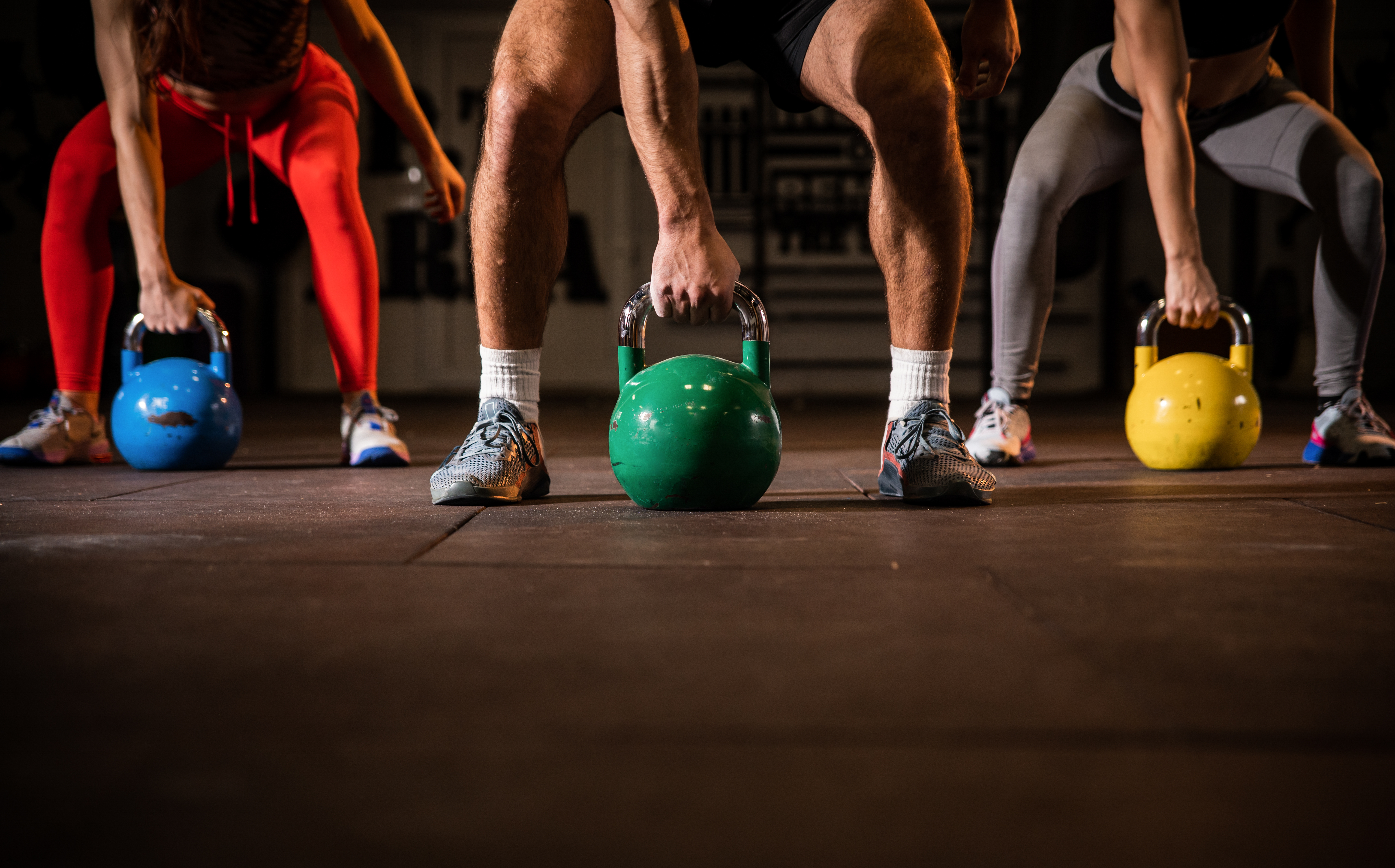 A low-angle shot of three people performing a kettlebell deadlift or swing, with a green, blue, and yellow kettlebell on the gym floor.