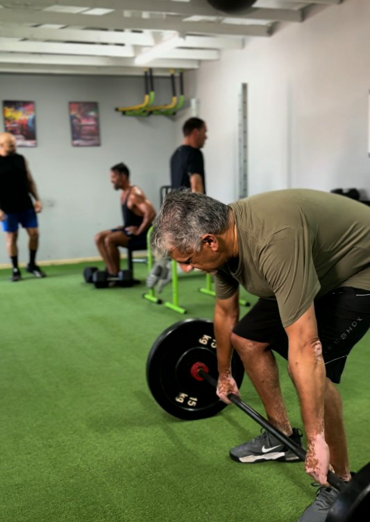 A man with grey hair, wearing an olive-green t-shirt, is performing a deadlift in a gym. He is gripping a barbell loaded with black weight plates, with other gym members visible in the background on a green turf floor.