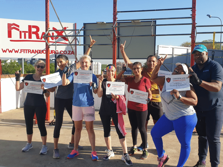 A group of individuals proudly displays their FitRanX Certificates of Completion after a successful outdoor training session. The photo celebrates their hard work and the sense of community in the program, with the FitRanX logo and website visible in the background.