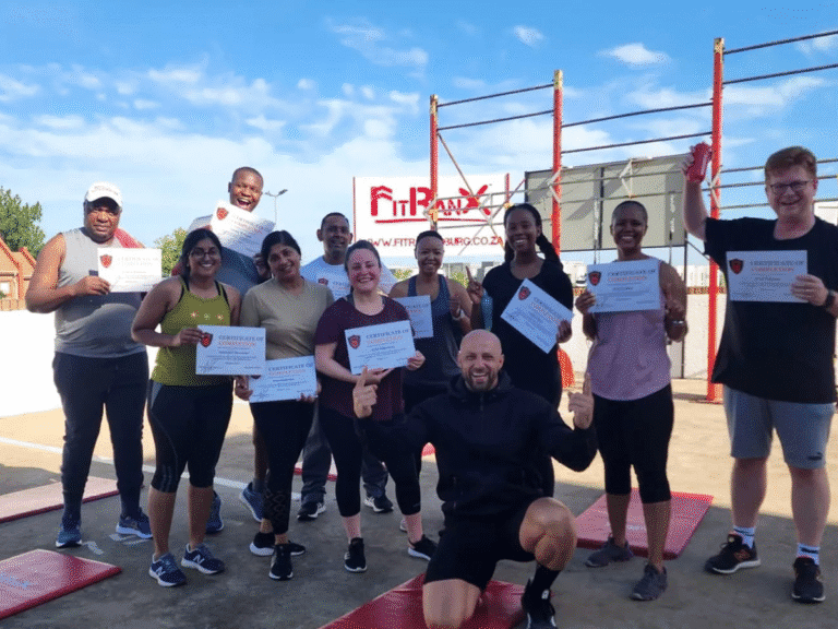 A group of nine people, including a coach kneeling in the center, are posing outdoors on a concrete training area. Each person is holding a FitRanX Certificate of Completion, with a large FitRanX sign and red training rig in the background.