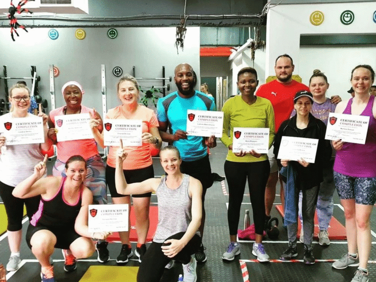 A group of people, dressed in various workout clothes, are posing for a group photo inside a gym. They are all holding up their FitRanX "CERTIFICATE OF COMPLETION," with some kneeling and others standing.