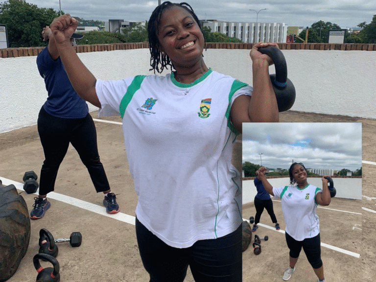 A smiling woman wearing a white and green sports shirt is posing triumphantly outdoors, flexing her bicep and holding a kettlebell. Other workout equipment and another person are visible in the background.