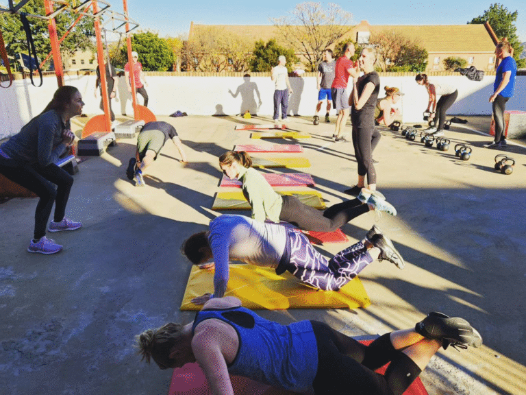 A diverse group of people is engaged in an outdoor workout. In the foreground, several individuals are on exercise mats doing push-ups. In the background, others are performing squats and working with kettlebells.