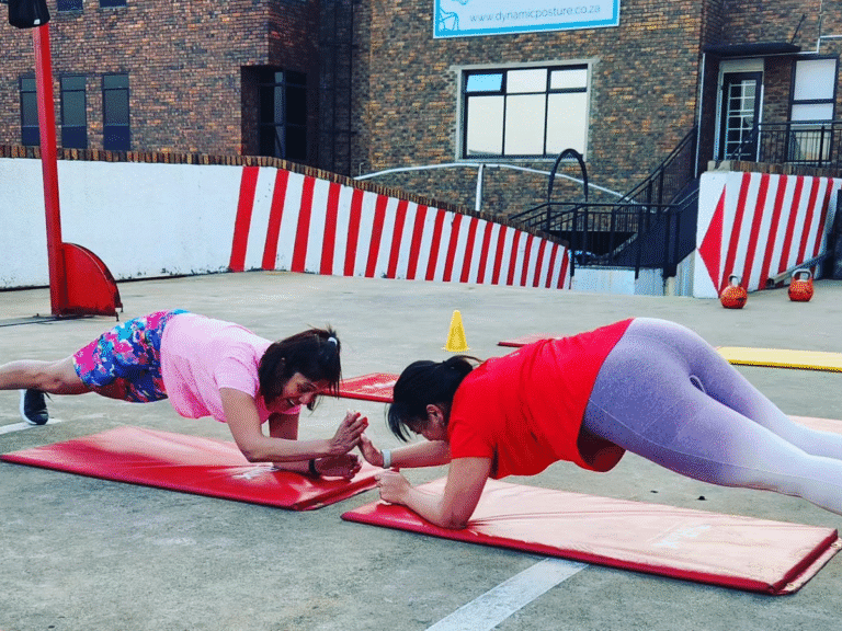 Two women in workout attire are doing a plank exercise on red mats outdoors. They are facing each other and giving a high-five, with a red and white striped wall in the background.