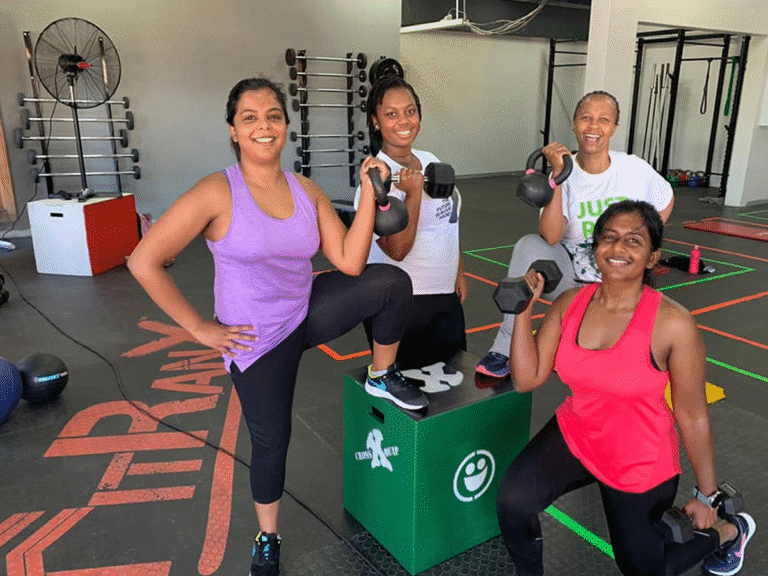 Four women in workout clothes are posing for a group photo in a gym. One woman is on a green box holding a kettlebell, another is holding a dumbbell, and the other two are smiling at the camera. The FitRanX logo is visible on the floor.
