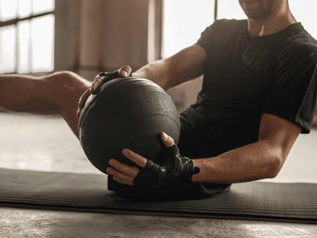 A man in a black t-shirt and gloves is sitting on a mat, holding a heavy black medicine ball while performing a core exercise. He is leaning back with his knees bent and feet off the ground, twisting his torso.